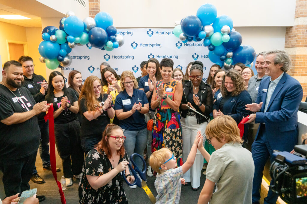 Group of individuals in front of a Heartspring logo backdrop clapping and celebrating the grand opening of the feeding therapy room