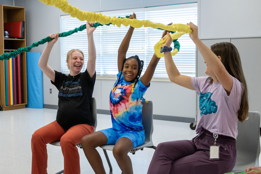 Child sitting between two Heartspring staff members in a group music therapy CARE Club session looking at the camera