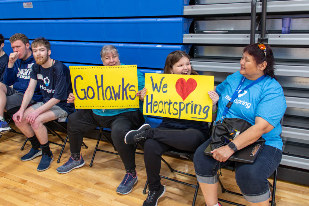Heartspring direct support professionals and students sitting and holding up "Go Hawks" and "We Heart Heartspring" signs at Special Olympics