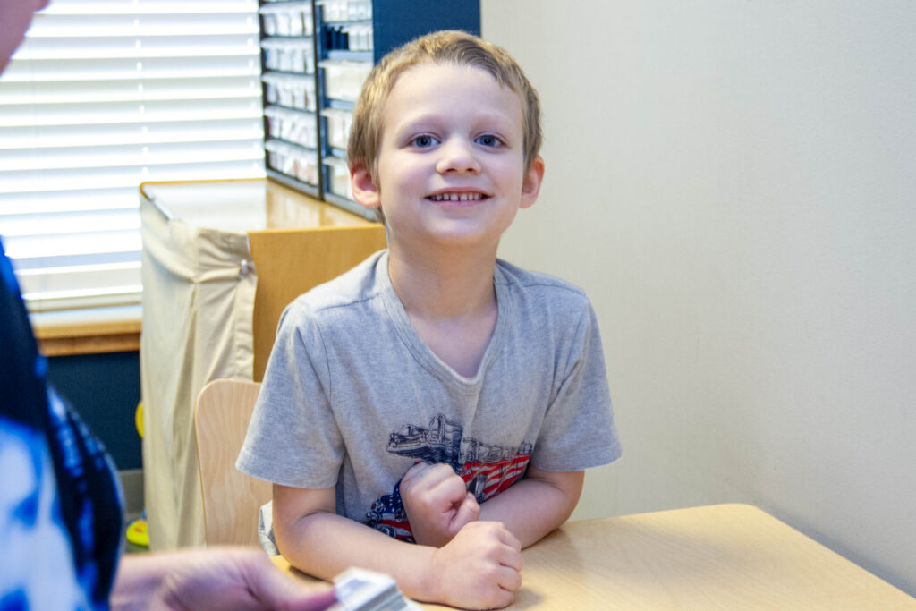 Julian sitting at a table and smiling at the camera during a pediatric speech-language therapy appointment