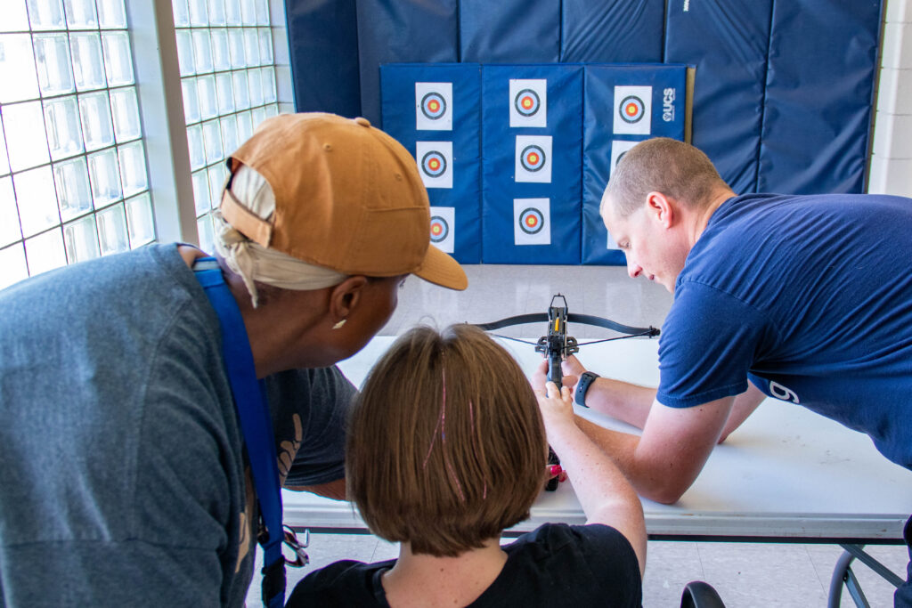 Special education paraprofessional and adpated physical education (APE) coach help a student aim at an archery target in P.E. class.