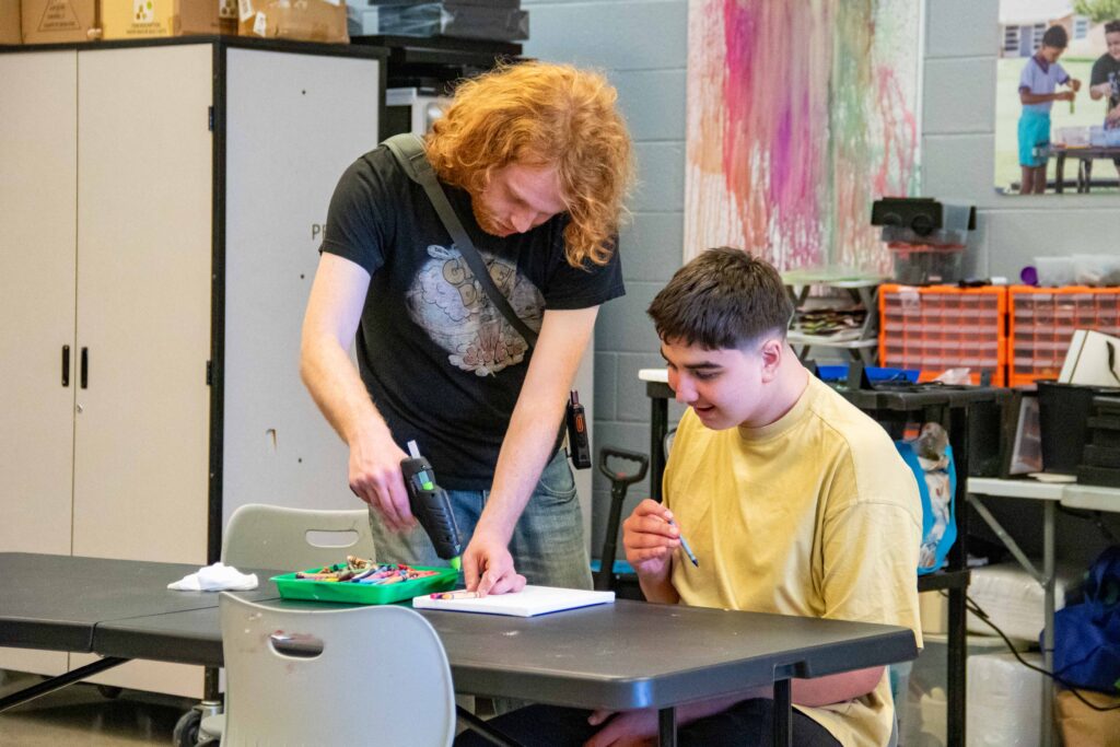 A special education paraprofessional helps a student melt crayons over a canvas in art class.