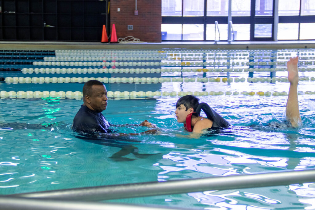 Adapted physical education (APE) coach helps a student kick their legs and they work on learning to swim in a pool.