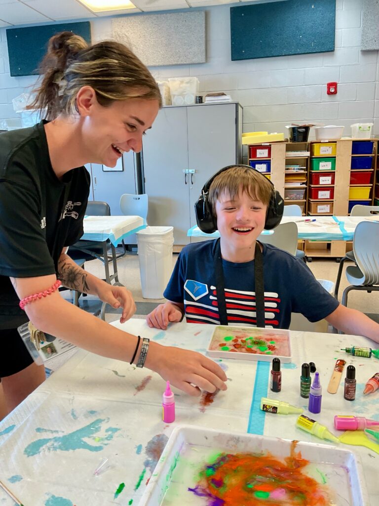 Heartspring Therapeutic Residential & Day School student and special education paraprofessional smile and make sand art together in art class.