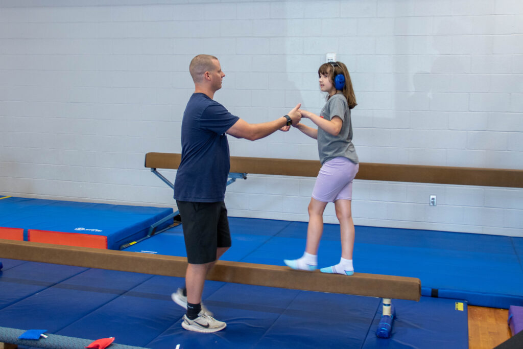 Adapted physical education (APE) coach helps a student balance as she crosses the balance beam in the YMCA adaptive gymnastics class.