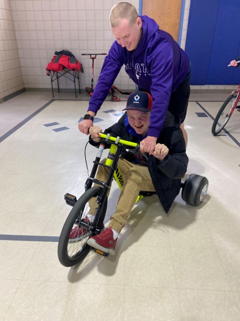 Student rides a tricycle with the assistance of the adapted physical education (APE) coach.