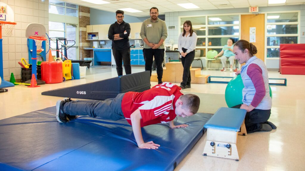 Brandon, a Heartspring Pediatric Outpatient Clinic client, works on his physical therapy skills doing a push-up with as his PT cheers him on and the WSU students observe.