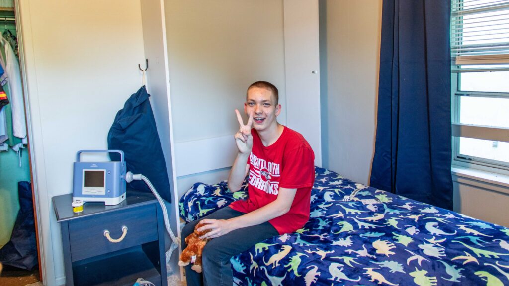 Brandon, a Heartspring Pediatric Outpatient Clinic client, enjoys his new Murphy bed solution and gives a peace sign to the camera, thanks to engineering students at Wichita State University.