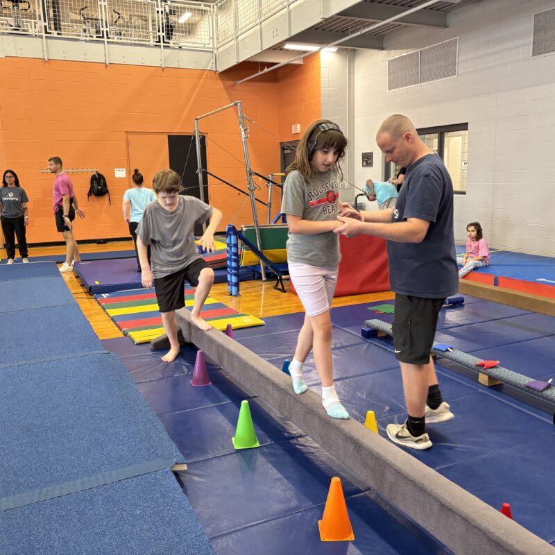 Adapted physical education (APE) teacher helps a student across the balance beam in adptive gymnastics at the YMCA.