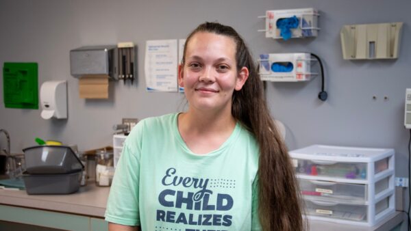 CMA, Karena Sweeten, smiles for the camera in the medical office in front of glove boxes on the wall and a plastic tray.