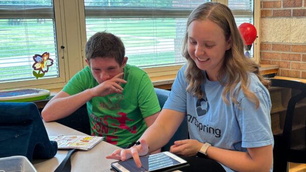 Taylor Lipford, SLP, works with a student in the Heartspring Therapeutic School at hyis desk. She is smiling and looking down as she enters data into a tablet.