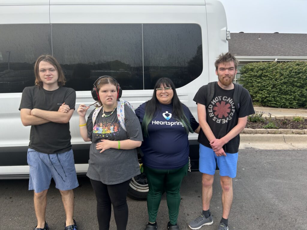 Direct Support Professional, Sareny Martinez, smiles for the camera alongside three Heartspring Therapeutic School students in front of a vehicle.
